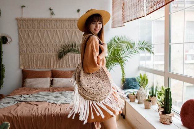 Lovely woman in linen dress and straw hat posing in boho style apartment