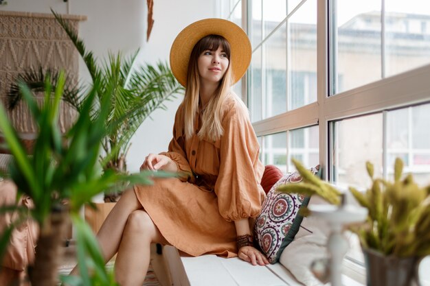 Lovely woman in linen dress and straw hat posing in boho style apartment