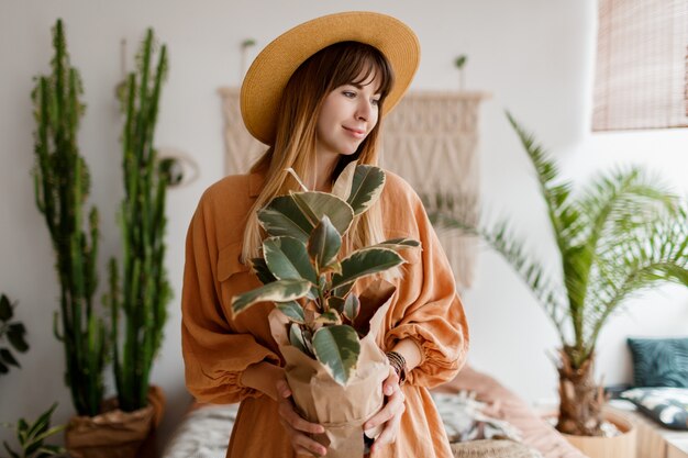 Милая женщина в льняном платье и соломенной шляпе позирует в квартире в стиле бохо