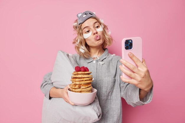 Lovely woman pouts lips and poses at front camera of smartphone poses with delicious pancakes for breakfast wears slumber suit holds soft pillow applies beauty patches poses against pink wall