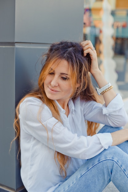 Lovely woman sitting on ground looking down and smiling in street during daytime .