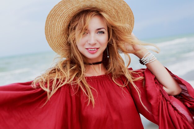 Красивая женщина в соломенной шляпе и модном красном летнем платье позирует возле океанских волн. Откровенная улыбка.