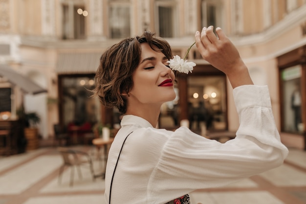 Lovely woman in white shirt with long sleeve holding flower in city. Short-haired woman with red lipstick posing at street.