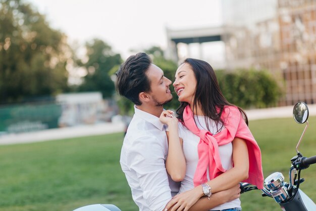 Lovely woman with black hair playfully kissing husband in good summer day on nature background