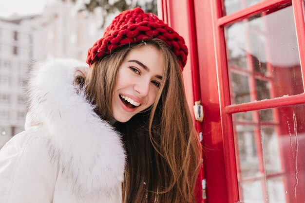 Прекрасная женщина с счастливой улыбкой позирует рядом с красной телефонной будкой в декабре утром. Открытый портрет замечательной европейской дамы зимой носит вязаную шапку и белое пальто.