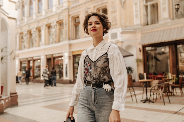 Lovely woman with wavy hair in jeans with belt and flower smiling at street. Cool lady in white blouse with lace posing in city.