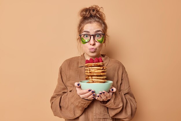 Lovely wondered woman keeps lips folded applies green hydrogel patches under eyes to reduce puffiness dressed in nightwear holds delicious pancakes with syrup has tasty breakfast applies beauty pads