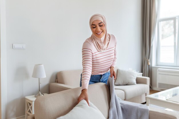 사랑스러운 젊은 아랍 이슬람 여성은 편안한 소파에 부드러운 베개와 격자 무늬를 넣어 집을 아늑하고 따뜻하게 만듭니다.