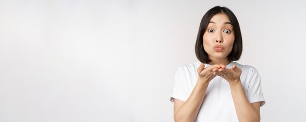 Lovely young asian woman sending air kiss and looking coquettish at camera standing in tshirt over w