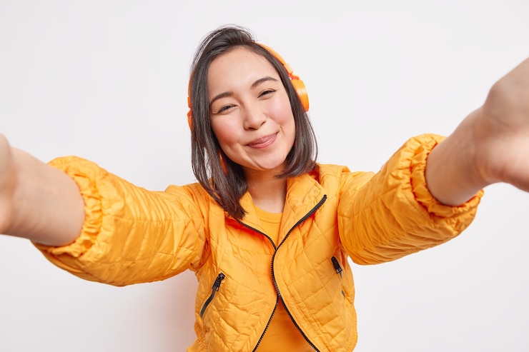 무료 Photo 검은 머리를 가진 사랑스러운 젊은 아시아 여성은 셀카 초상화 미소를 짓고 주황색 아노락 옷을 입고 음악을 듣고