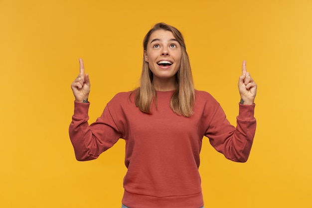 Lovely young beautiful woman smiles broadly and indicates with finger to copyspace posing with happy facial expression wears red pullover and blue denim pants