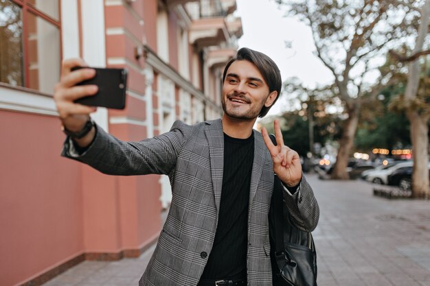 Lovely young boy with brunette hair and bristle, standing in black t-shirt and grey blazer, video chatting on phone and smiling, against city street