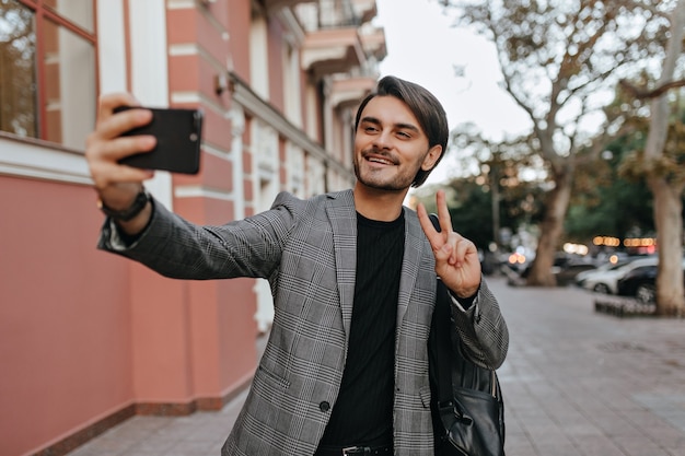 Lovely young boy with brunette hair and bristle, standing in black t-shirt and grey blazer, video chatting on phone and smiling, against city street