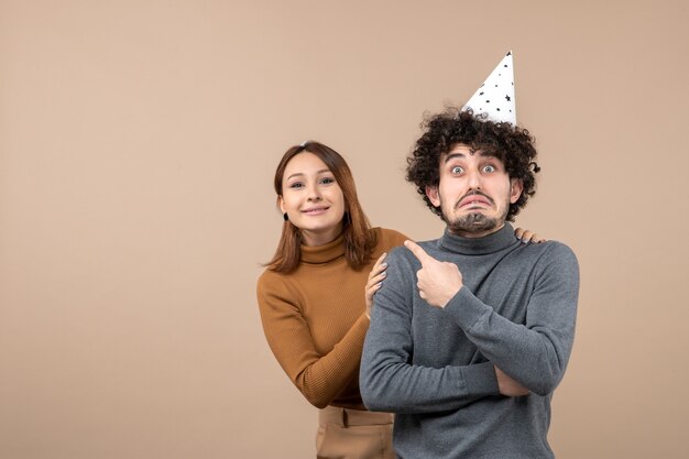 Lovely young couple wear new year hat angry girl looking at something standing behind upset guy pointing her on gray