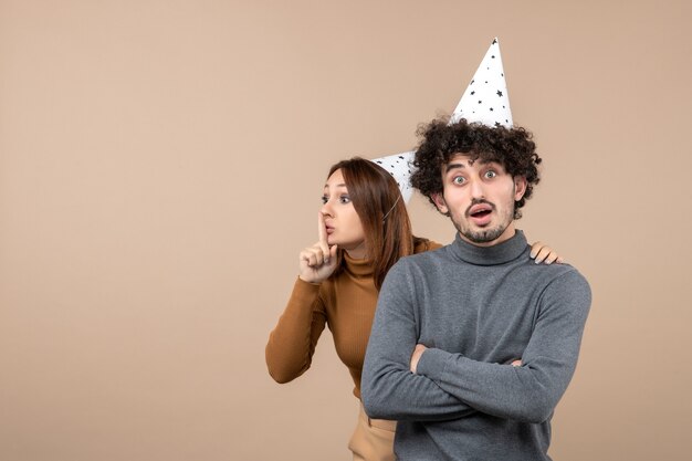 Lovely young couple wear new year hat girl making silence gesture to someone standing behind guy on gray