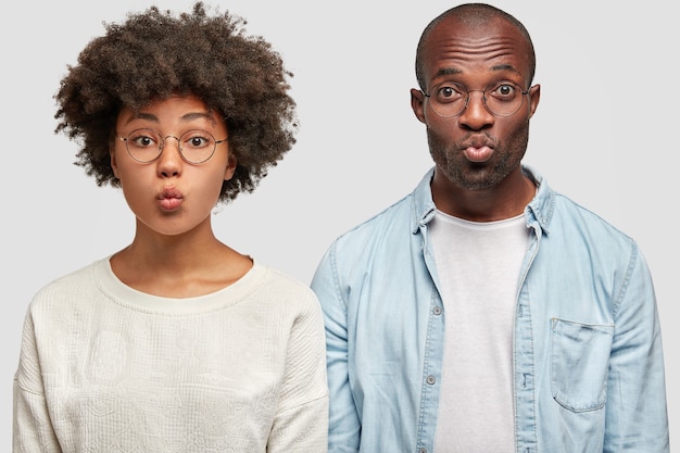 Lovely young dark skinned female has wavy hair, African American guy in denim shirt, stand next to each other, round lips, make grimace, isolated over white wall. Friendship concept