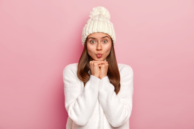 Lovely young female keeps lips folded, hands under chin, has appealing look into camera, wears white winter outfit, has healthy skin, well cared complexion, isolated over pink wall. Face expressions