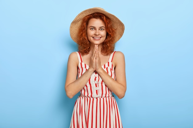 Lovely young female models indoor, keeps palms pressed together, being thankful for help, wears summer striped dress, straw hat,  isolated on blue wall. People, body language