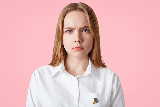 lovely young female with gloomy expression, frowns face, has long hair, healthy skin, blue eyes, white shirt, poses on pink. Discontent woman has quarrel with boyfriend