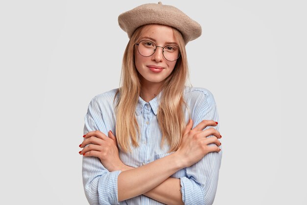 Lovely young French woman in stylish beret and shirt keeps arms folded, looks with pleased expression, happy to recieve compliment, isolated over whitewall. People and lifestyle concept