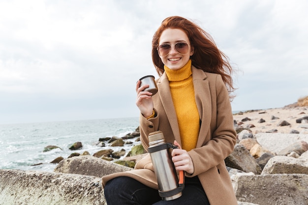 Милая молодая рыжая женщина в осеннем пальто гуляет по пляжу, пьет горячий кофе из термоса