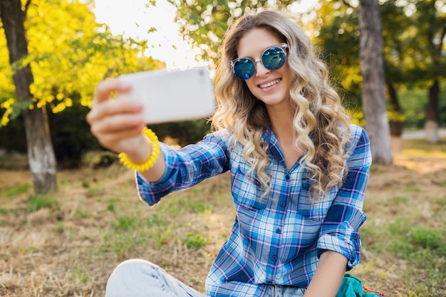 Lovely young stylish attractive smiling blond woman sitting in park, summer casual style