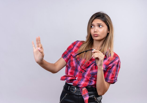 A lovely young woman in checked shirt holding sunglasses showing stop gesture on a white wall