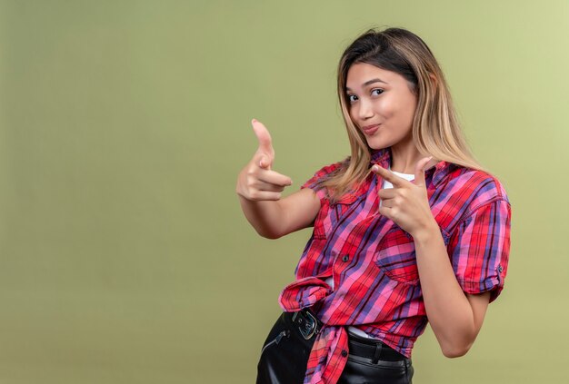 A lovely young woman in a checked shirt pointing with index fingers on a green wall