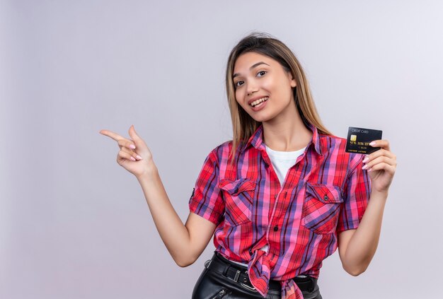 A lovely young woman in a checked shirt showing credit card and pointing with index finger 