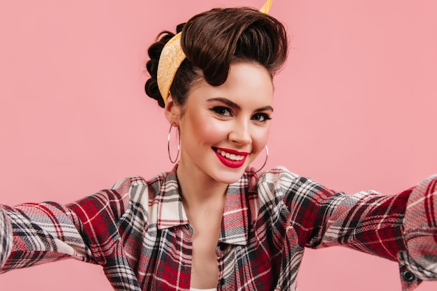 Free photo lovely young woman in checkered shirt smiling on pink background. emotional pinup girl taking selfie.