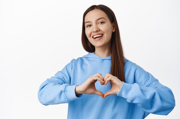 Lovely young woman in hoodie showing heart I love you sign, smiling and looking adorable, express her tender feelings, standing against white background
