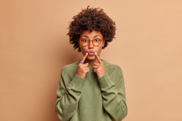 Lovely young woman keeps index fingers near folded lips makes face at front wants to kiss someone wears glasses and sweater isolated over brown wall