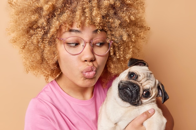 Free photo lovely young woman keeps lips folded holds pug dog wants to kiss favorite pet wears spectacles and casual pink t shirt isolated over beige background female pet lover enjoys company of friend