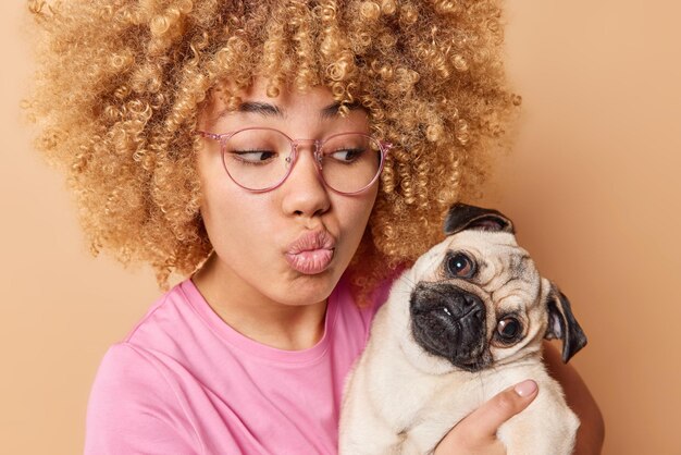 Lovely young woman keeps lips folded holds pug dog wants to kiss favorite pet wears spectacles and casual pink t shirt isolated over beige background Female pet lover enjoys company of friend