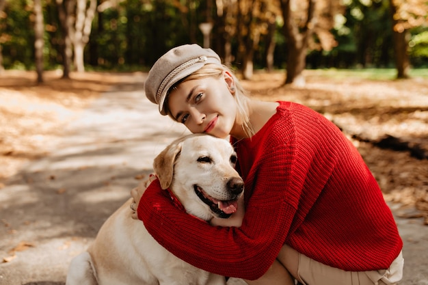 Прекрасная молодая женщина в красивой светлой шляпе и красном свитере, сидя с лабрадором вместе в осеннем парке. Симпатичная блондинка и ее собака сидят среди опавших листьев.