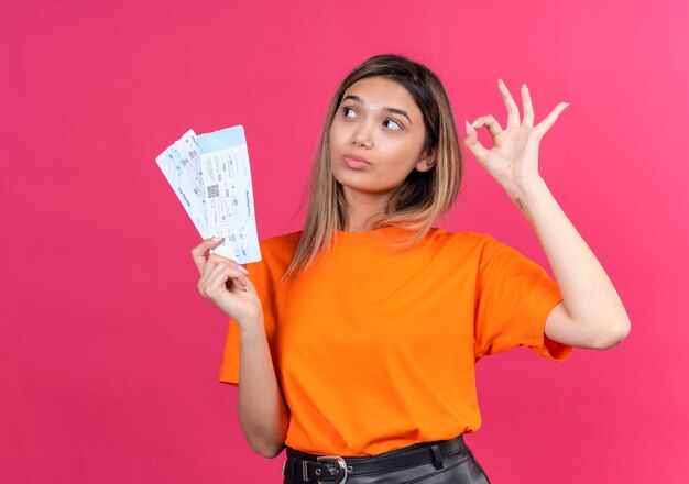 A lovely young woman in an orange t-shirt showing ok gesture while holding plane tickets on a pink wall