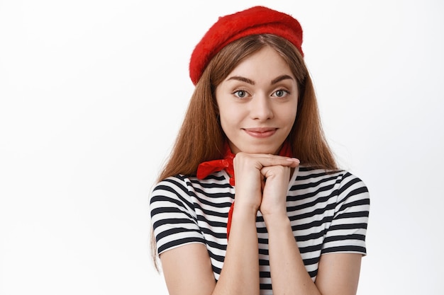 Lovely young woman in red beret, gazing at front, smiling and looking at you with admiration and interest, standing over white wall