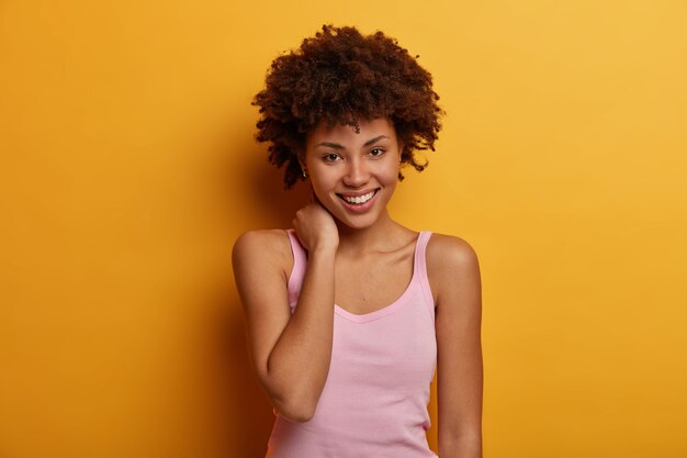 Lovely young woman touches neck, looks with tender smile, feels good and touched, wears casual t shirt, has direct gaze , isolated on yellow wall. Positive human face expressions