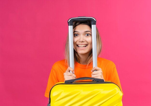Милая молодая женщина в оранжевой рубашке, улыбаясь и глядя, держит желтый чемодан на розовой стене