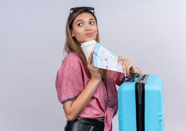 A lovely young woman wearing red shirt in sunglasses looking side while holding blue suitcase and plane tickets on a white wall