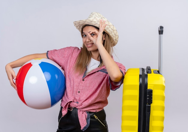 A lovely young woman wearing red shirt and sunhat holding inflatable ball and put hand on yellow suitcase while showing ok gesture on a white wall