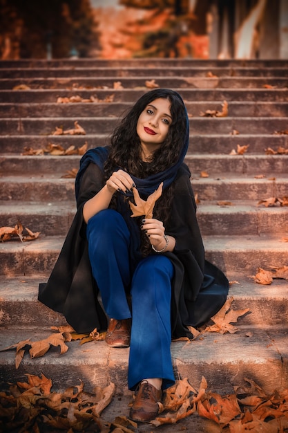 Lovely young woman wears knitted scarf and black dress, sitting on the stairs in autumn park.