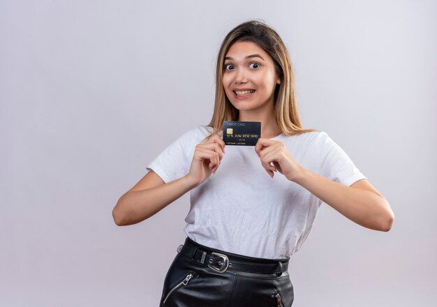Милая молодая женщина в белой футболке улыбается, показывая кредитную карту на белой стене