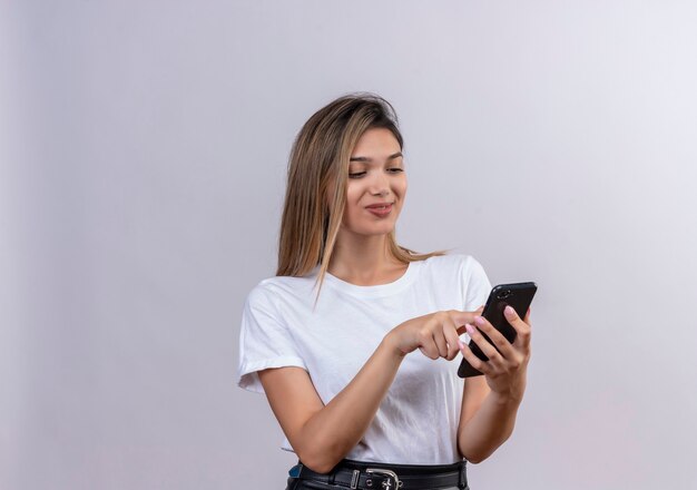 A lovely young woman in white t-shirt smiling while touching screen of mobile phone on a white wall