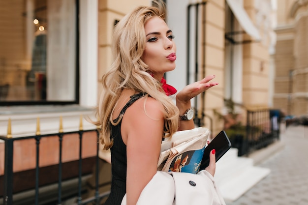 Lovely young woman with curly hairstyle sends air kiss to someone standing on the street