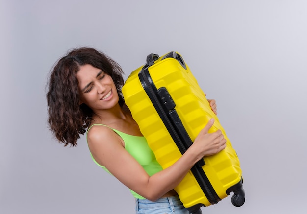 A lovely young woman with short hair in green crop top carrying heavy yellow suitcase on a white background