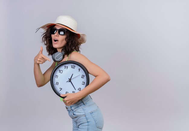 A lovely young woman with short hair in green crop top wearing sunglasses and sun hat pointing at wall clock with index finger on a white background