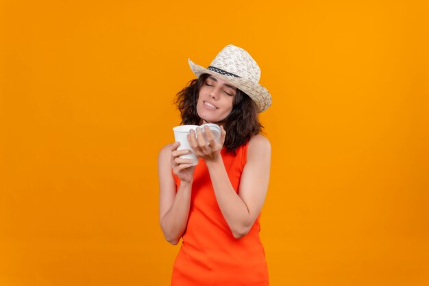 A lovely young woman with short hair in an orange shirt wearing sun hat enjoying the smell of coffee 