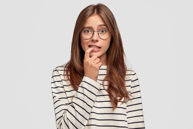 Lovely young woman with thoughtful expression bites finger, dressed in striped sweater, wears round spectacles, poses against white wall. Pensive worried female student thinks about something