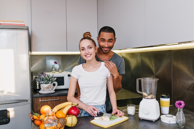 Lovers are cooking together in the kitchen. Girl with fair hair cuts fruits. Couple in T-shirts with joyful faces spend time together at home.
